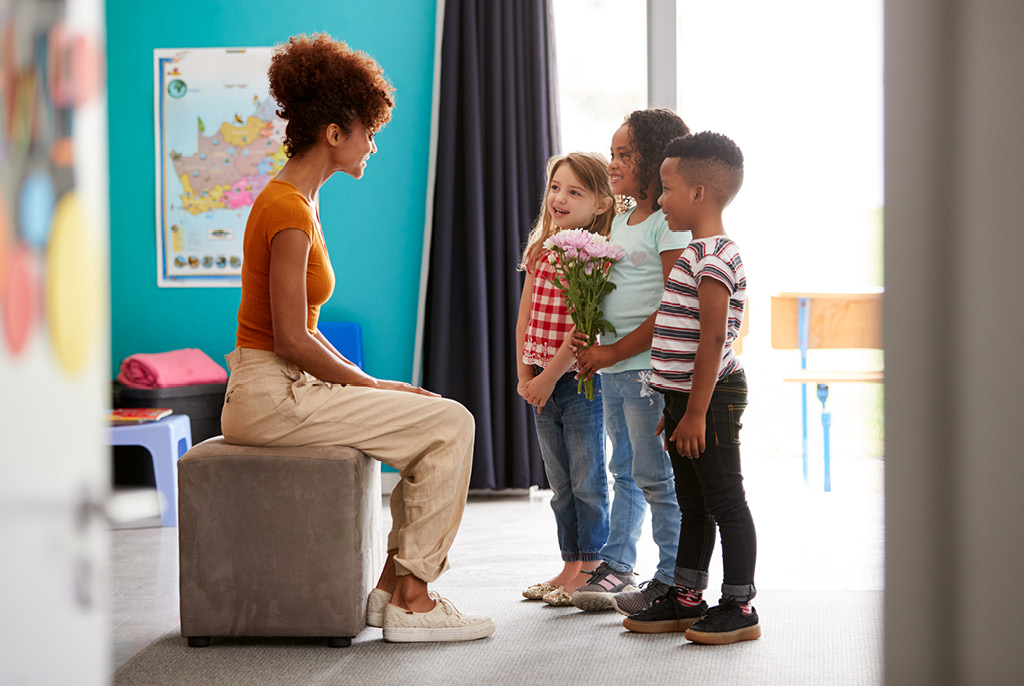 A group of children stand before their teacher with a bouquet of pink and white flowers.