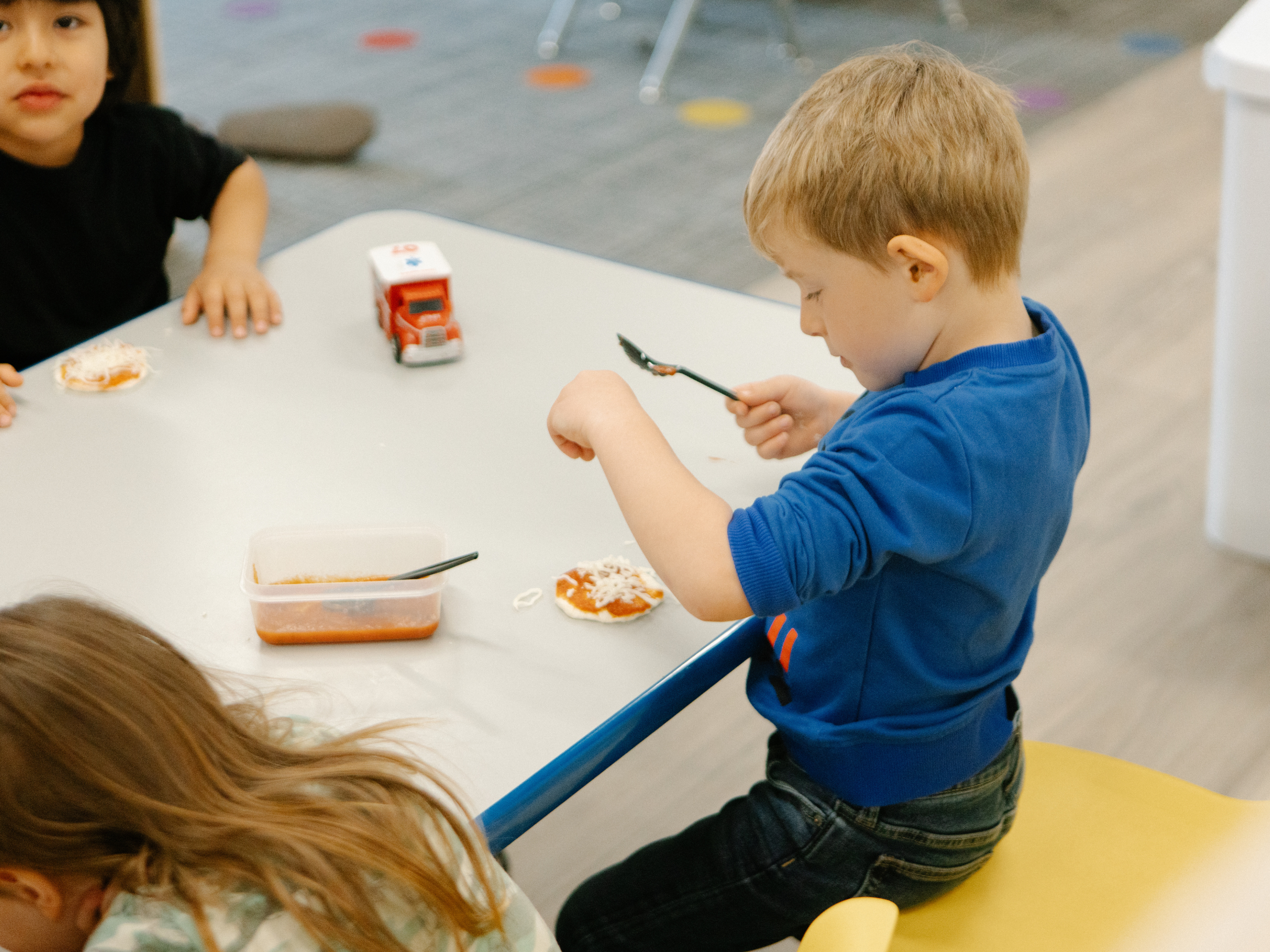 Children in an early education setting assemble mini pizzas during a hands-on activity.