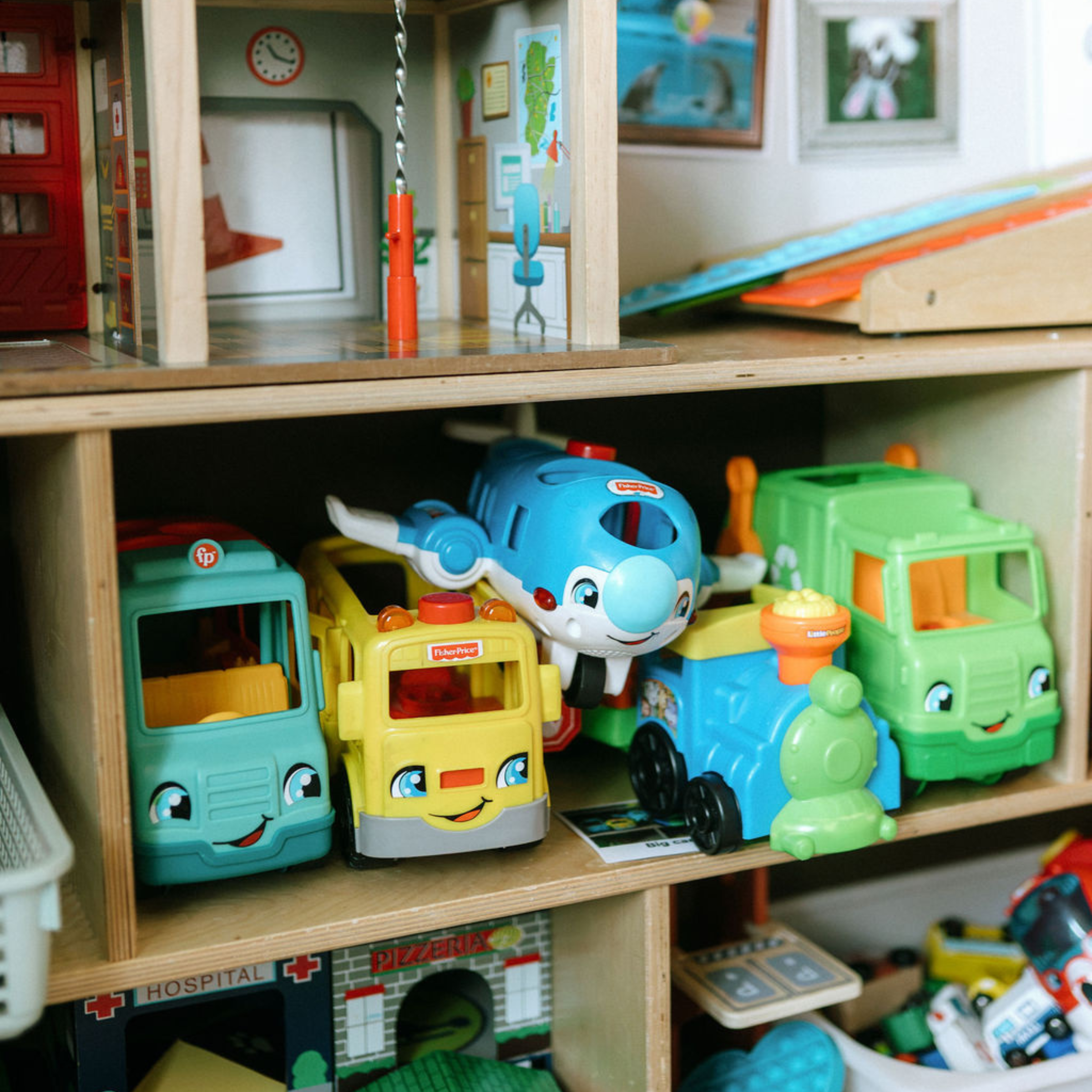 En una estantería de juguetes del aula se exponen diversos juguetes de colores, entre los que se incluyen autobuses de juguete, camiones, un tren, un avión y una casa de muñecas de madera.